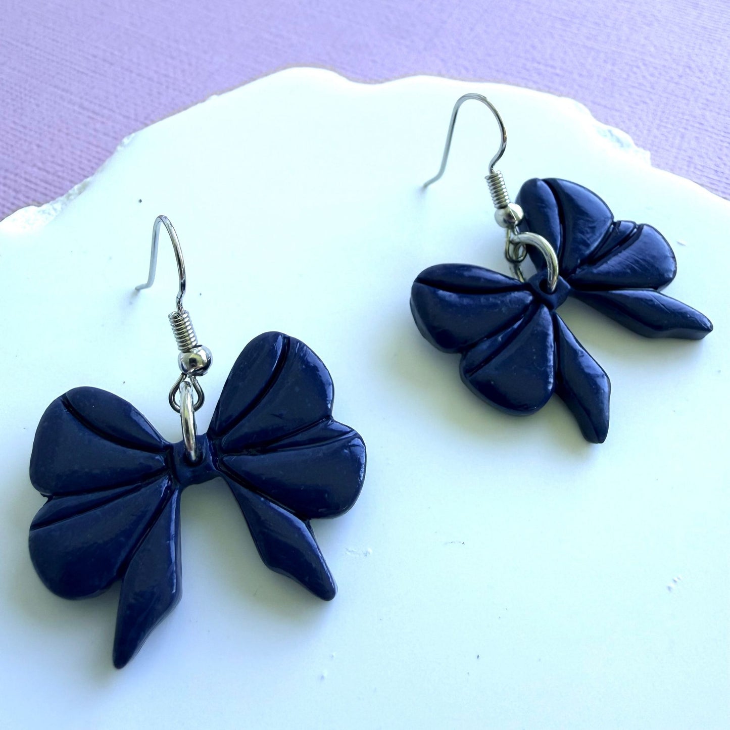 Navy blue bow-shaped handmade dangle earrings with silver hooks on a white surface with a light purple background.