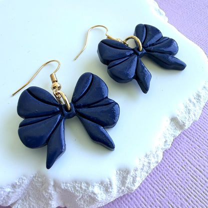 Navy blue bow-shaped dangle earrings with gold toned hooks on a white and purple textured surface.