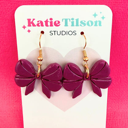 Purple bow-shaped dangle earrings on a Katie Tilson Studios card against a pink background.