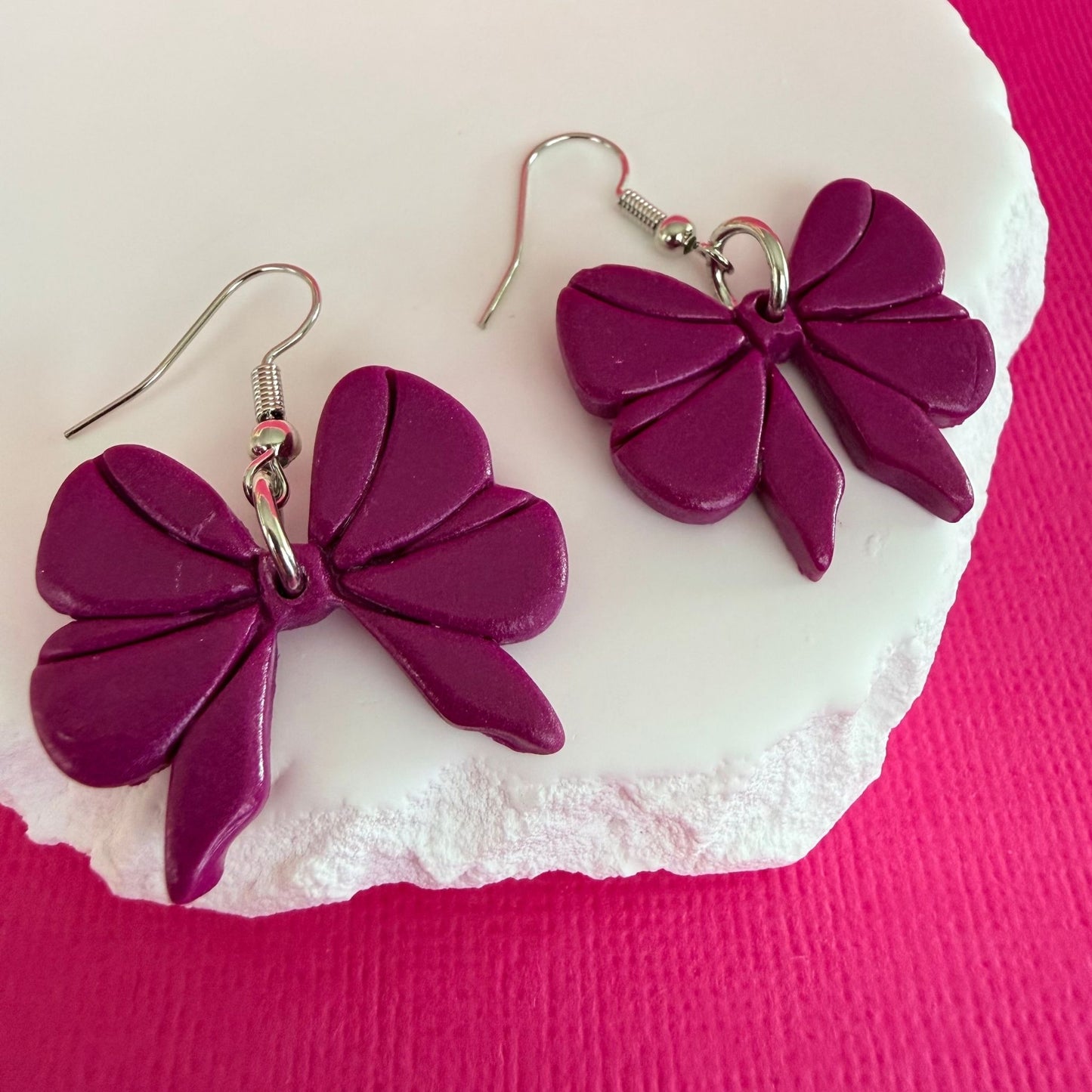 Australian handmade purple bow-shaped dangle earrings with silver toned hooks on a white stone with a pink background.
