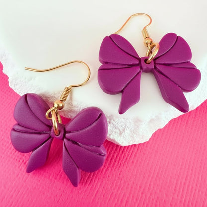 Purple bow-shaped handmade dangle earrings with gold hooks on a pink and white background.