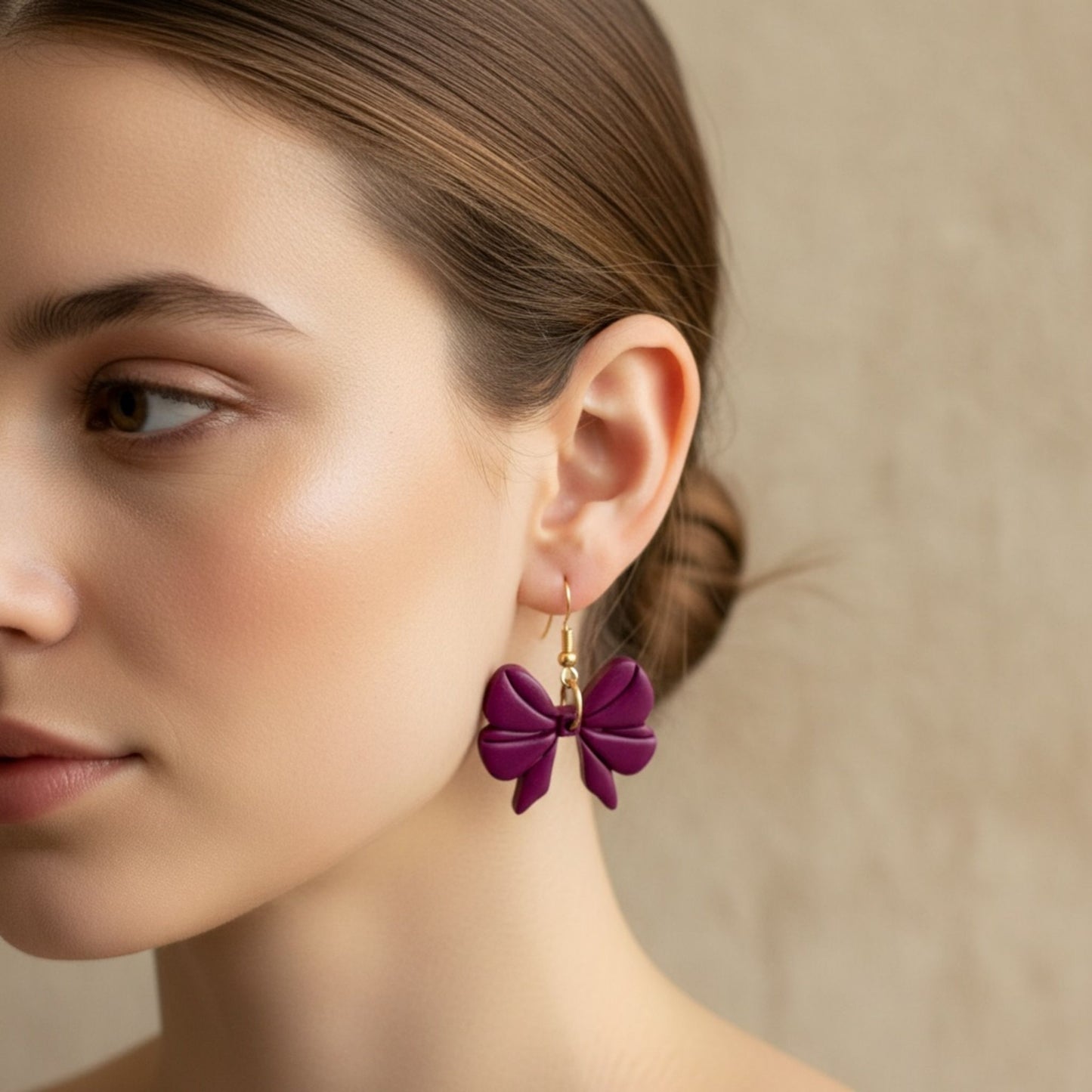 Close-up of a woman wearing purple bow-shaped dangle earrings with gold toned hooks against a neutral background.