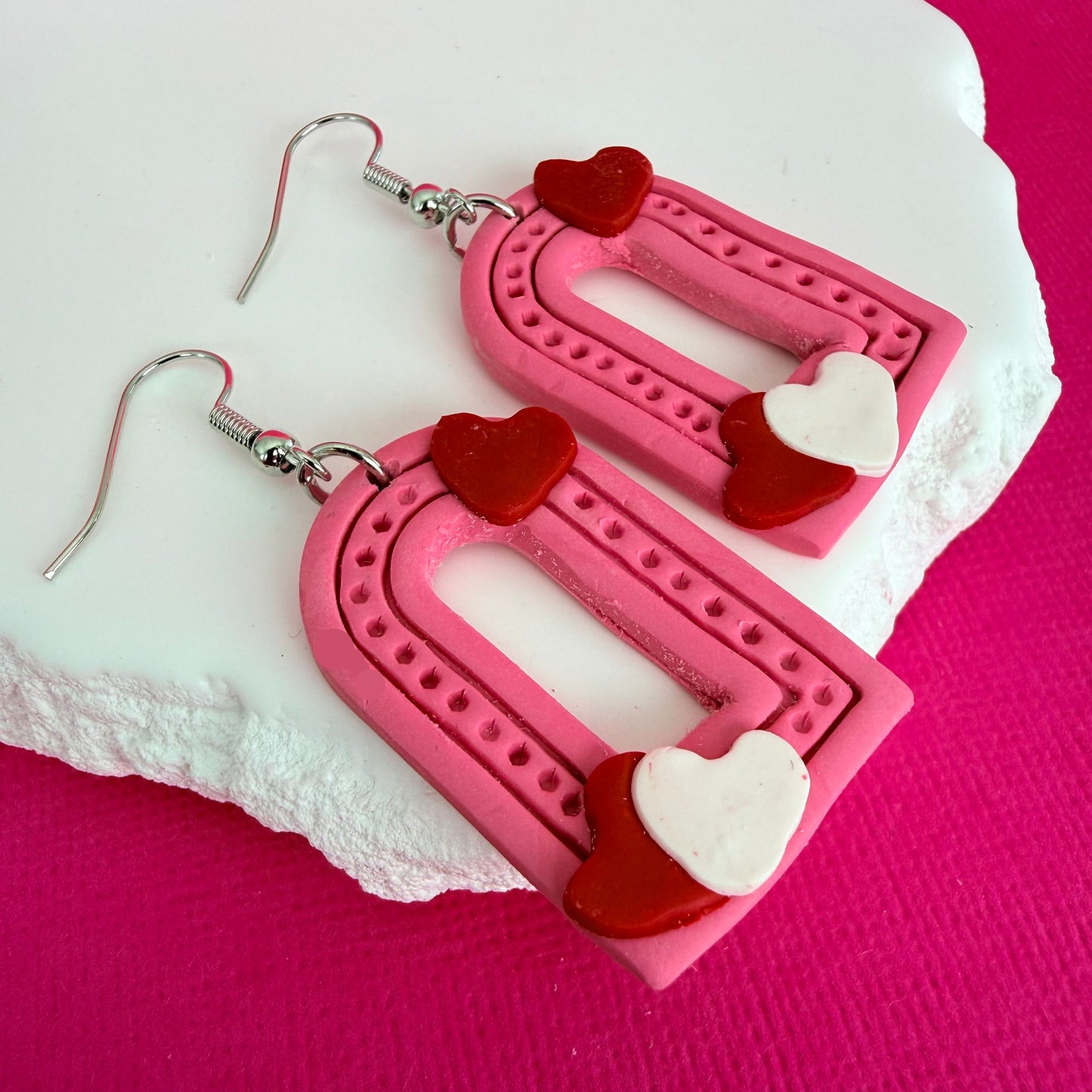 Australian handmade pink arch shaped earrings with red and white heart details and silver toned hooks on a pink background.