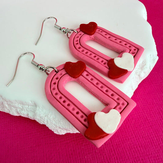 Australian handmade pink arch shaped earrings with red and white heart details and silver toned hooks on a pink background.