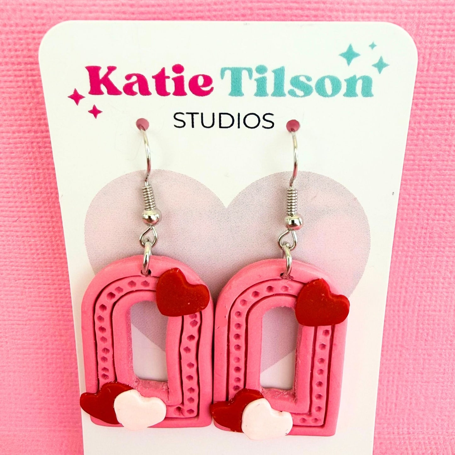 Love Note Arches handmade dangle earrings in pink arch design with red and white heart details on a Katie Tilson Studios branded earring card against a pink background.