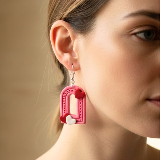 Close-up of a woman wearing pink arched shaped dangle earrings with heart details against a neutral background.