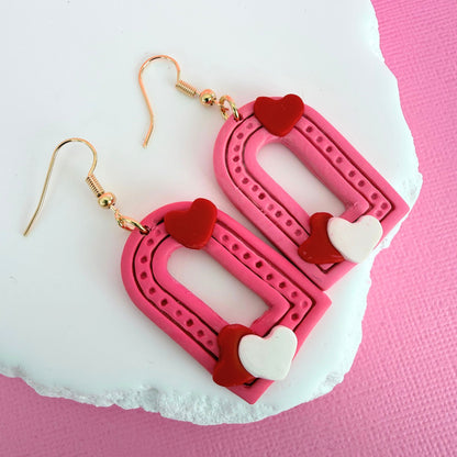 Pink arched shaped dangle earrings with heart details and gold hooks on a pink background.