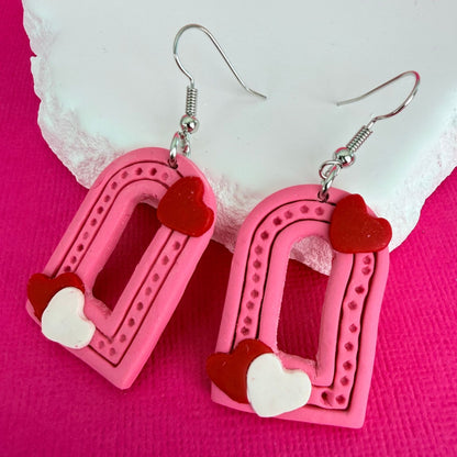 Pink heart-shaped dangle earrings with red and white heart details and silver toned hooks on a pink background.