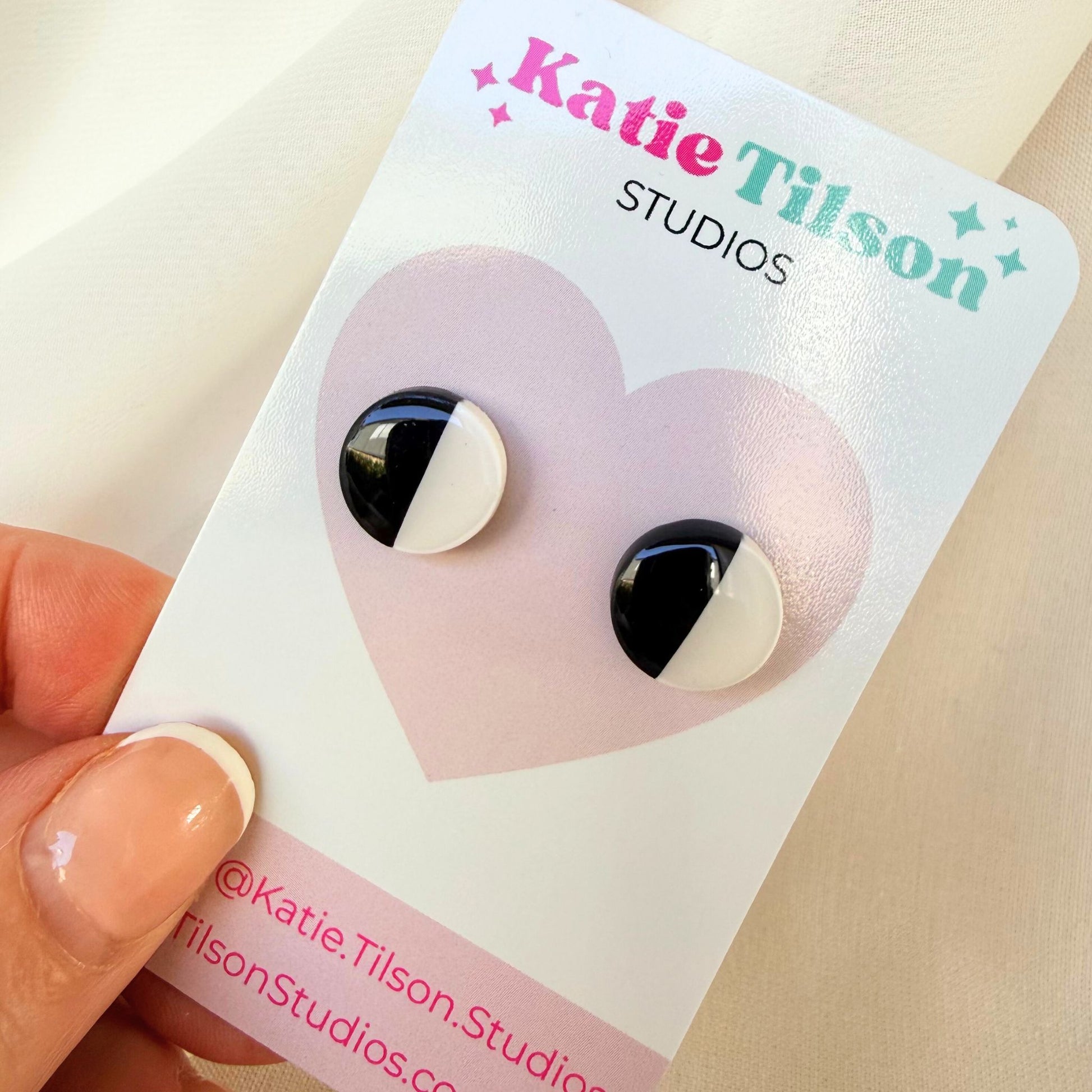 Black and white half and half circular stud earrings on a Katie Tilson Studios earring card held by a hand.