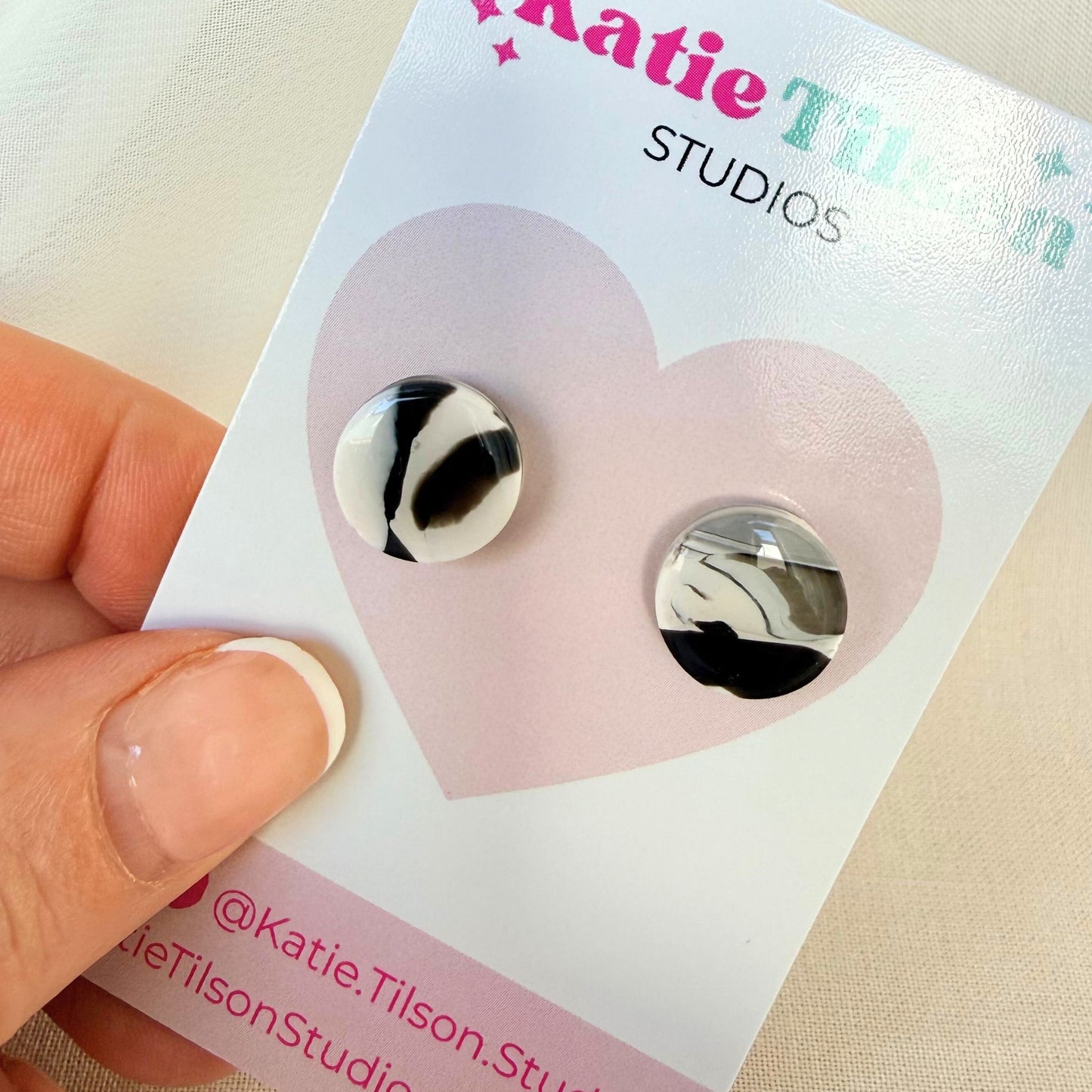 Midnight black and white marbled design circular stud earrings on a Katie Tilson Studios earring card held by a hand.