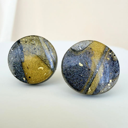 A pair of gold, silver and graphite coloured marbled circle studs with shimmer and glossy resin finish resting on a white surface.