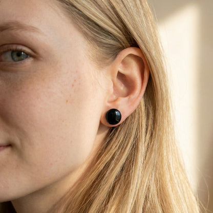 Close-up of a woman wearing midnight black stud earrings with a glossy finish against a blurred background.