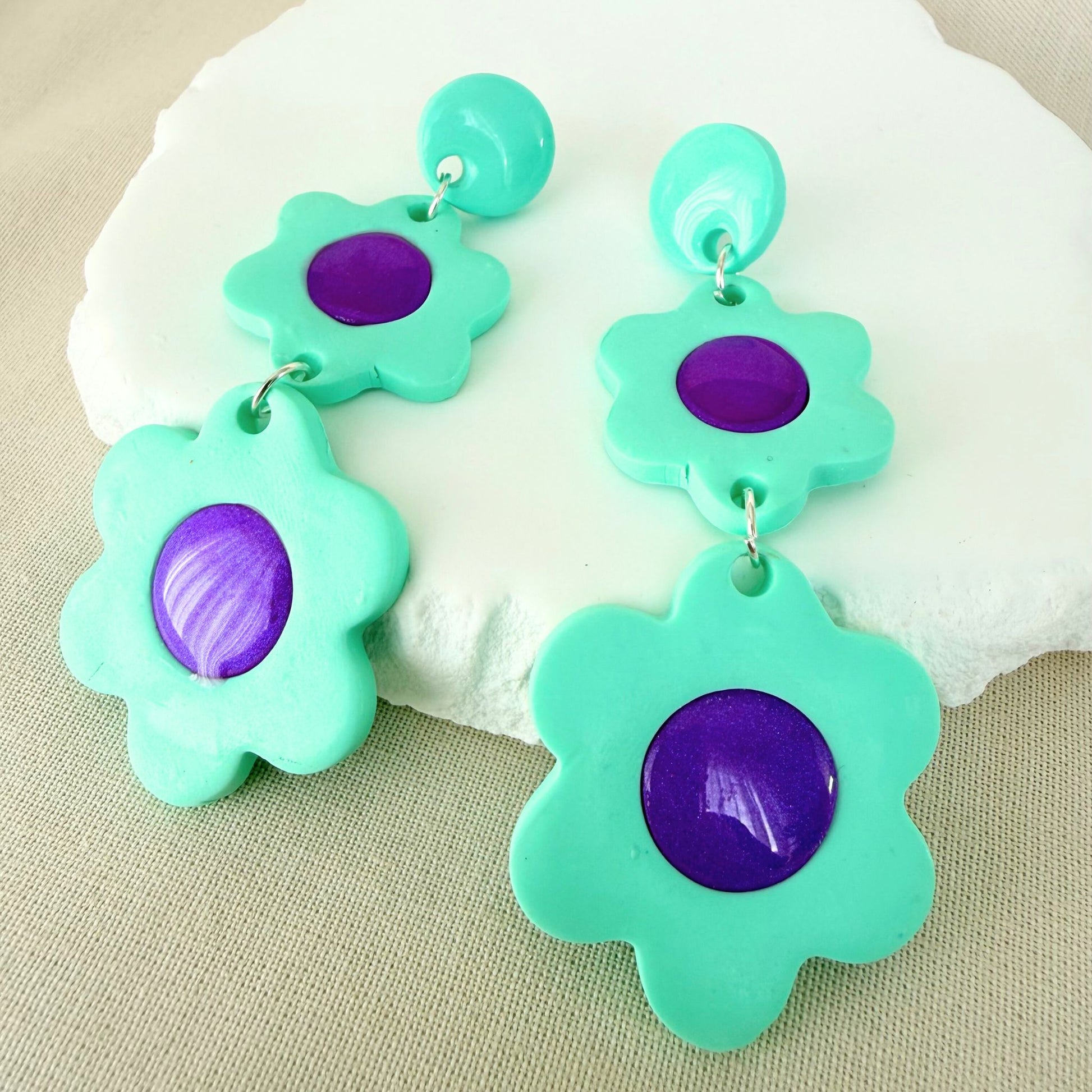Mint green flower dangle earrings with glossy violet centres on a white surface.