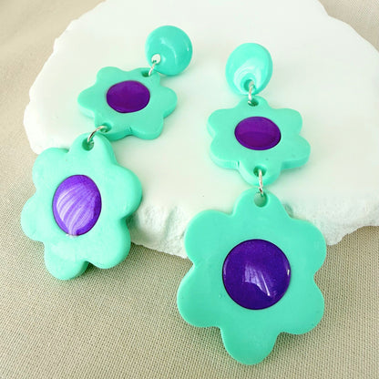 Mint green flower dangle earrings with glossy violet centres on a white surface.