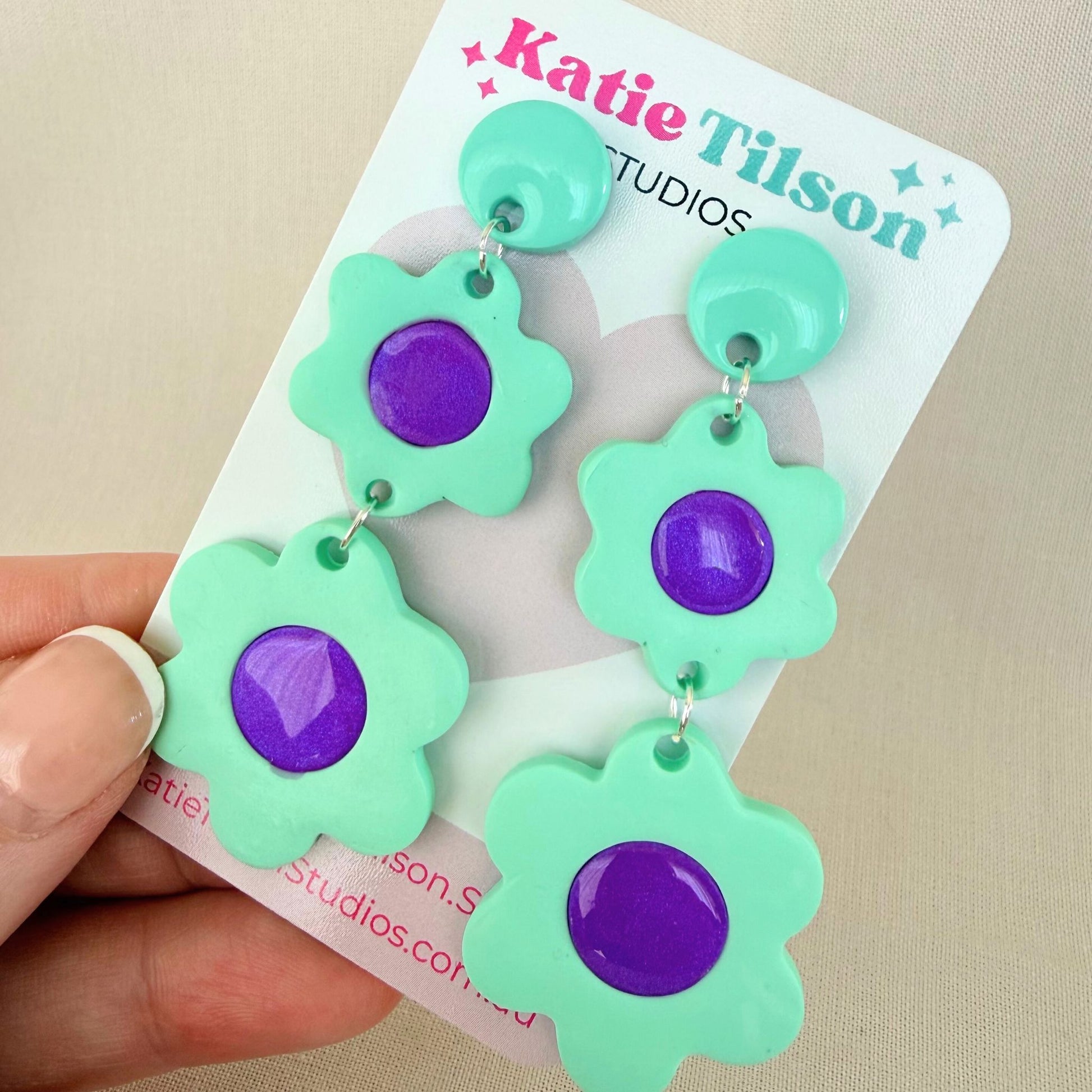 Mint green and purple floral dangle earrings on a Katie Tilson Studios card held by a hand.