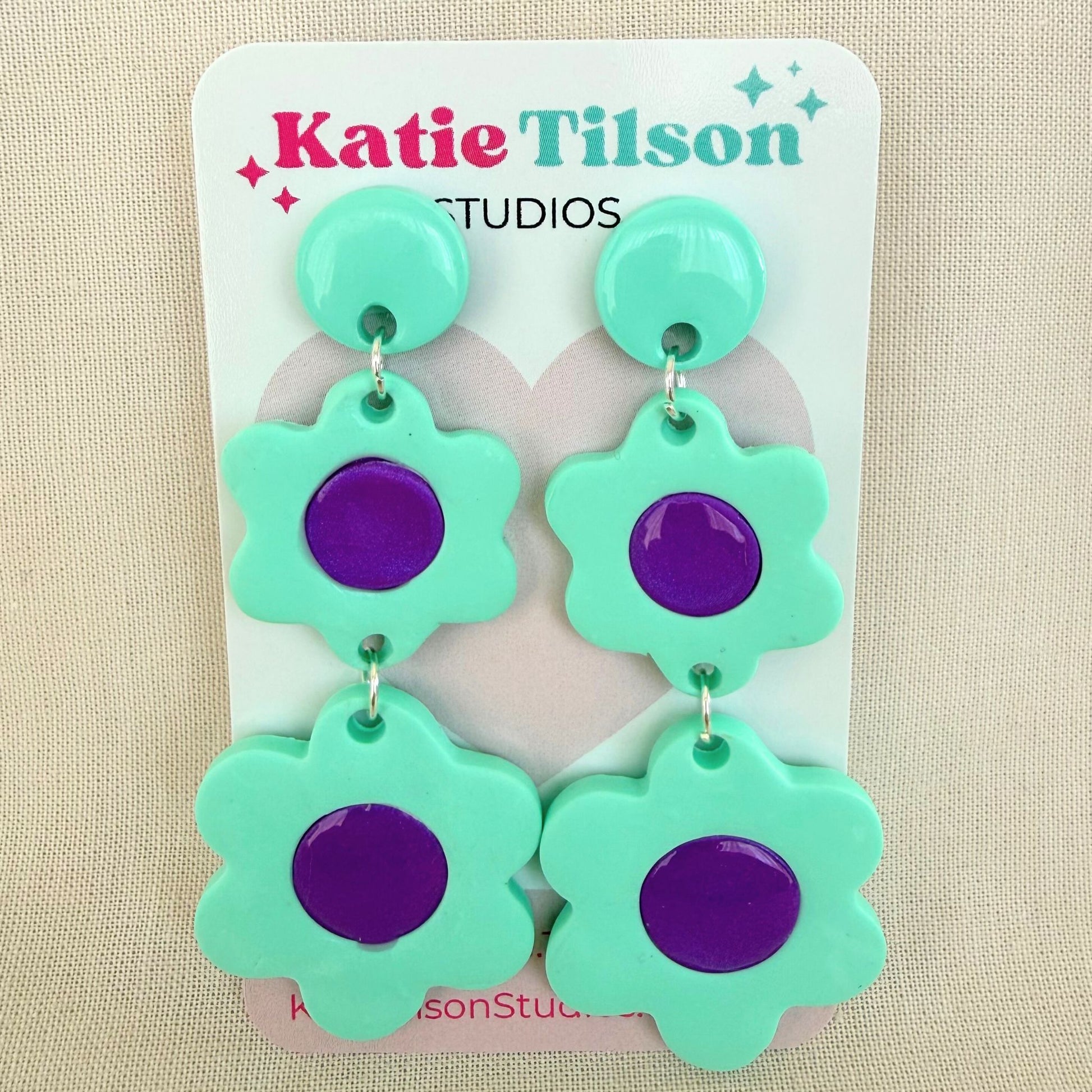 Green and purple flower-shaped dangle earrings on a Katie Tilson Studios card against a beige background.