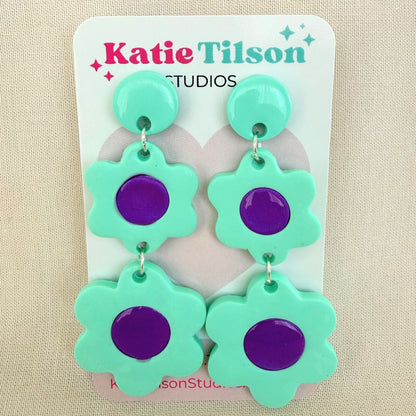 Green and purple flower-shaped dangle earrings on a Katie Tilson Studios card against a beige background.