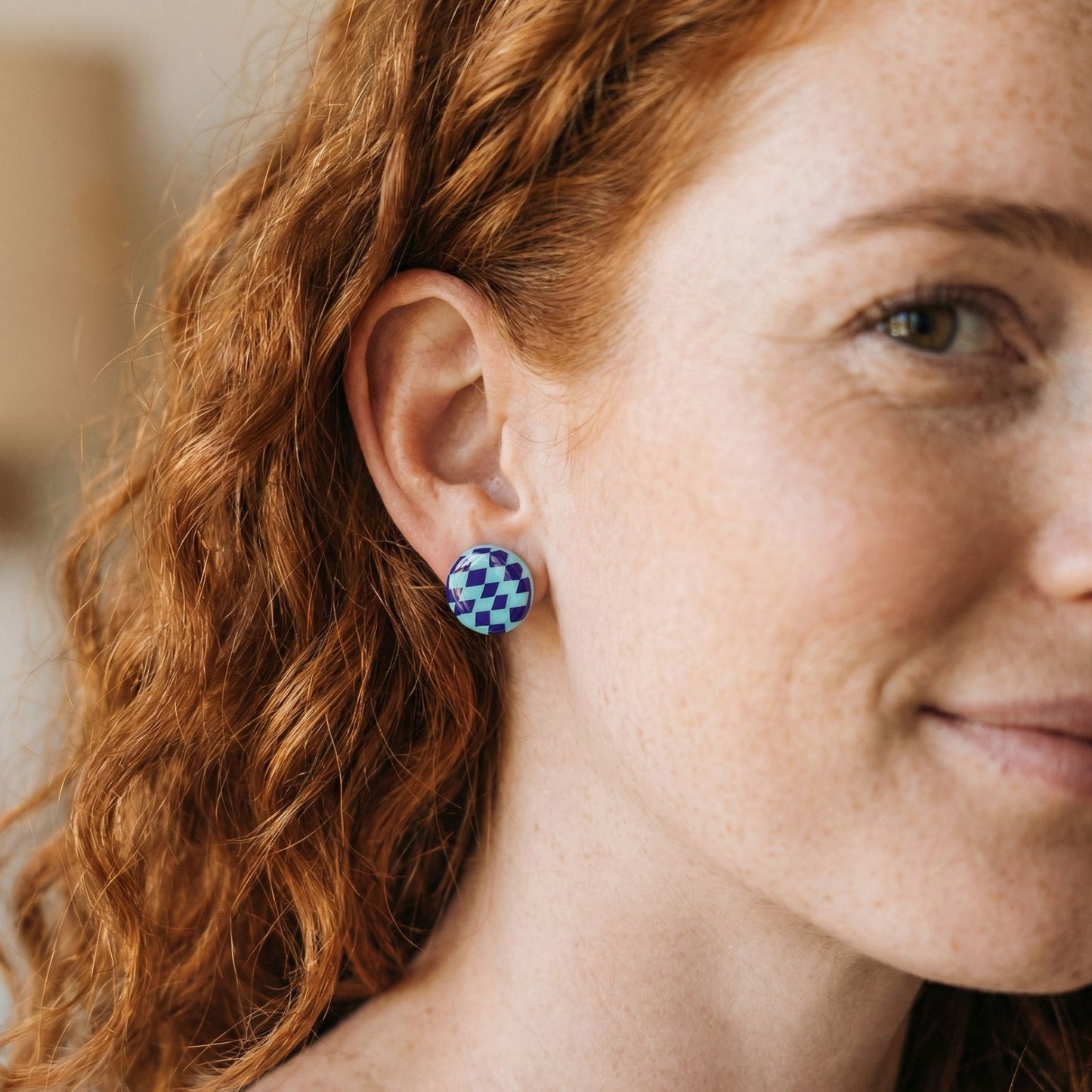 Close-up of a woman wearing a mint and purple checkered stud earring with a blurred background.
