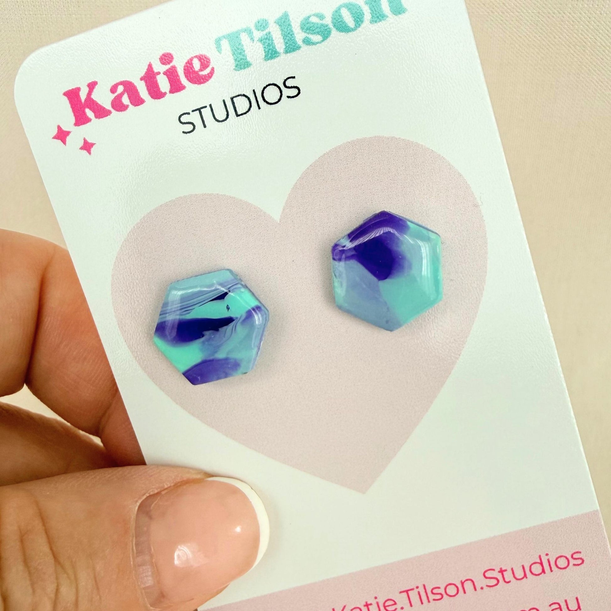 Violet and mint geometric stud earrings on a Katie Tilson Studios card held by a hand.