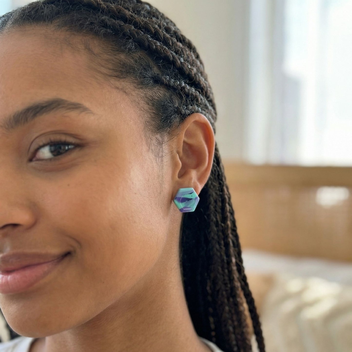 Close-up of a woman wearing colorful violet and mint hexagon shaped stud earrings with a blurred indoor background.