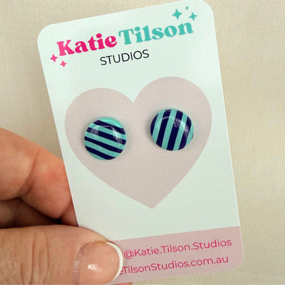 Mint and violet circular shaped striped stud earrings on a Katie Tilson Studios card held by a hand.
