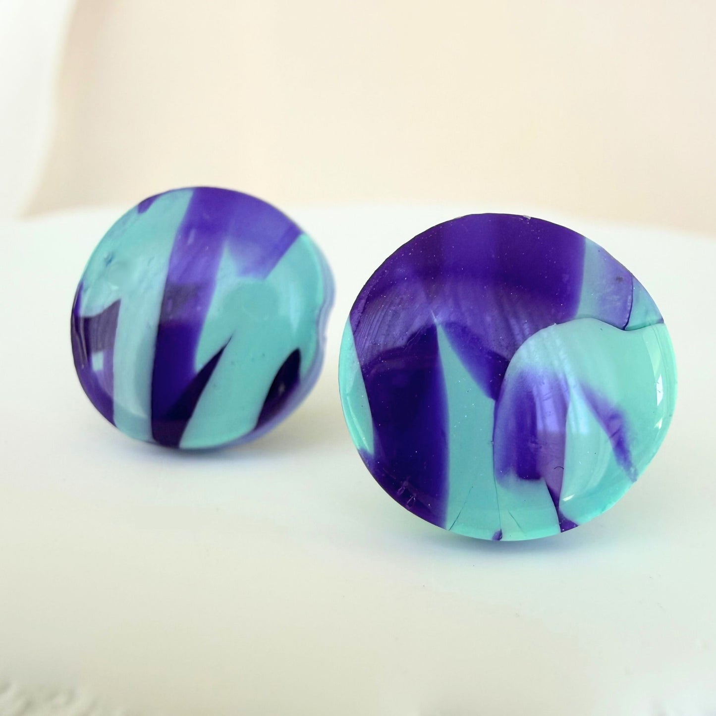 A pair of violet purple and mint abstract design circle studs with a glossy resin finish resting on a white surface.