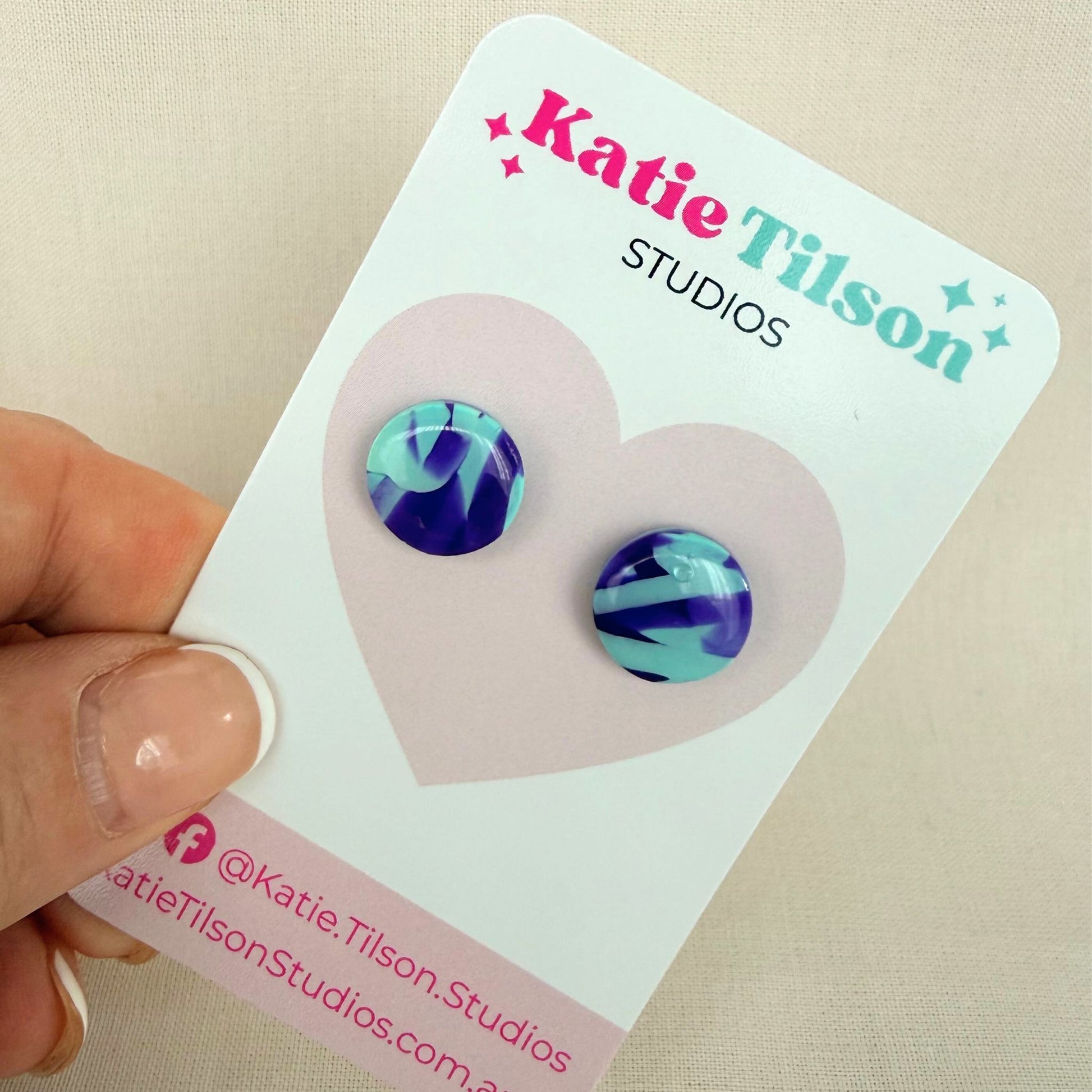 Mint and violet abstract design circular stud earrings on a branded earring card being held by a hand.