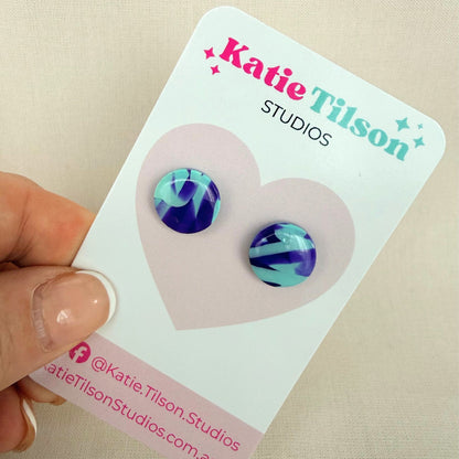 Mint and violet abstract design circular stud earrings on a branded earring card being held by a hand.