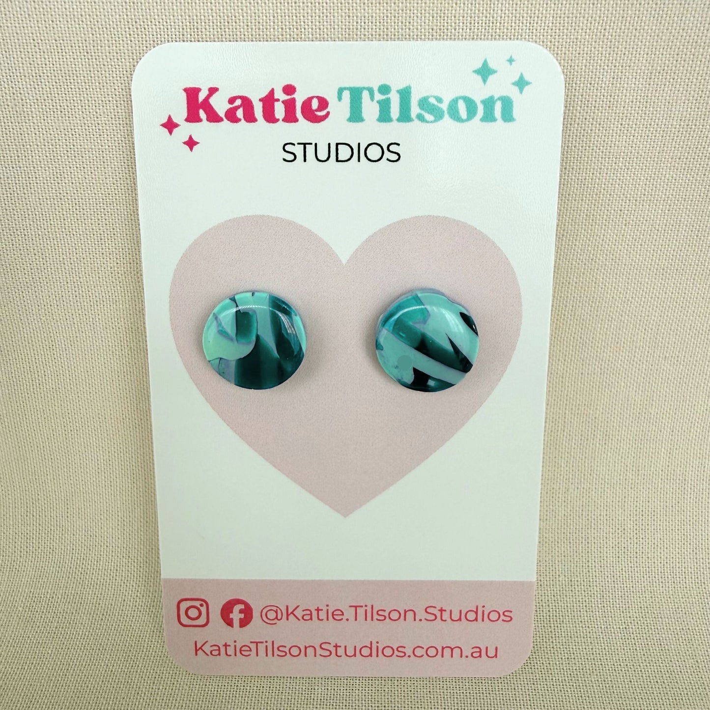 Mint and violet abstract design circular stud earrings on a branded earring card against a beige background.