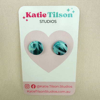 Mint and violet abstract design circular stud earrings on a branded earring card against a beige background.
