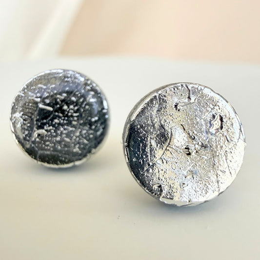 A pair of silver circle studs with mirror shine and a glossy resin finish resting on a white surface.