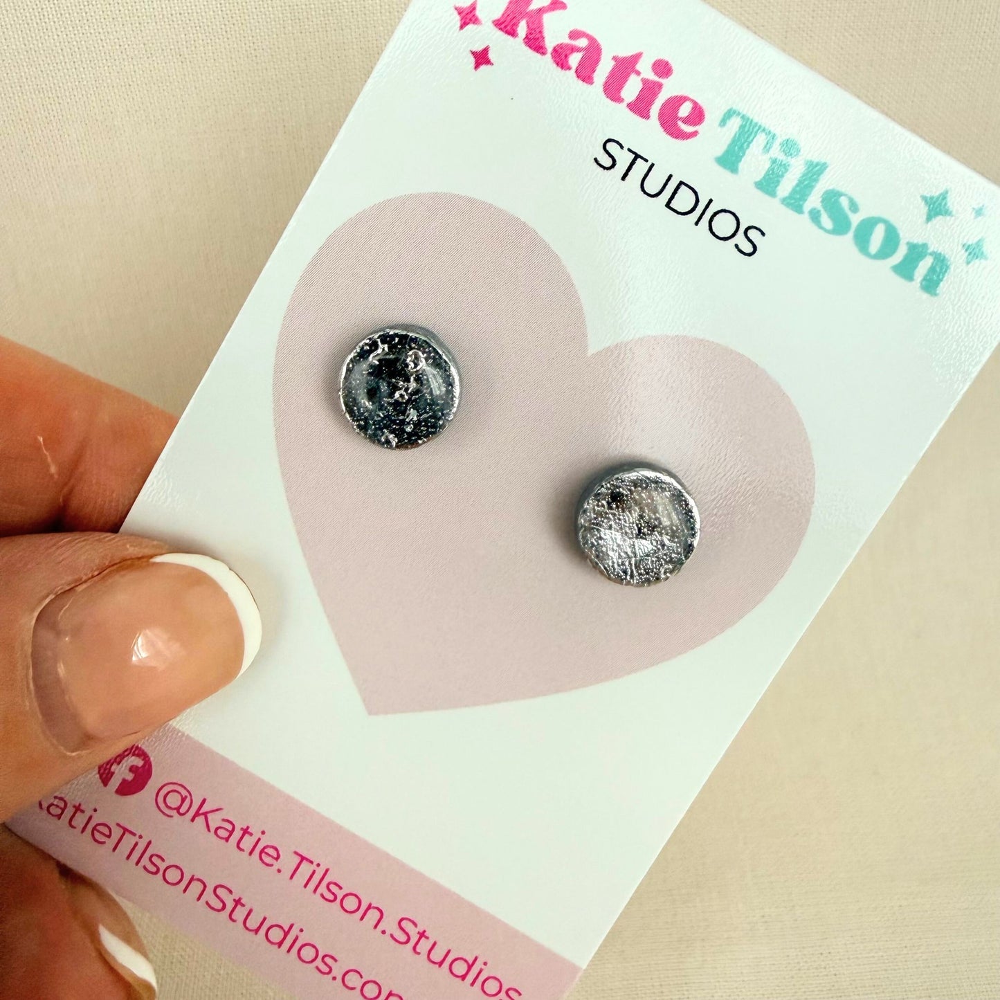 Pair of silver coloured circular stud earrings on a Katie Tilson Studios card held by a hand.