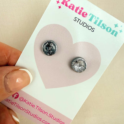 Pair of silver coloured circular stud earrings on a Katie Tilson Studios card held by a hand.