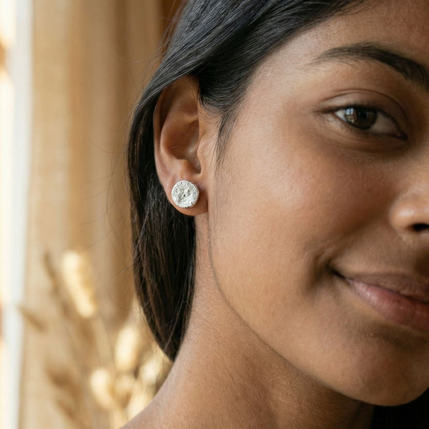 Close-up of a woman wearing round silver reflective stud earrings with a glossy finish against a blurred background.