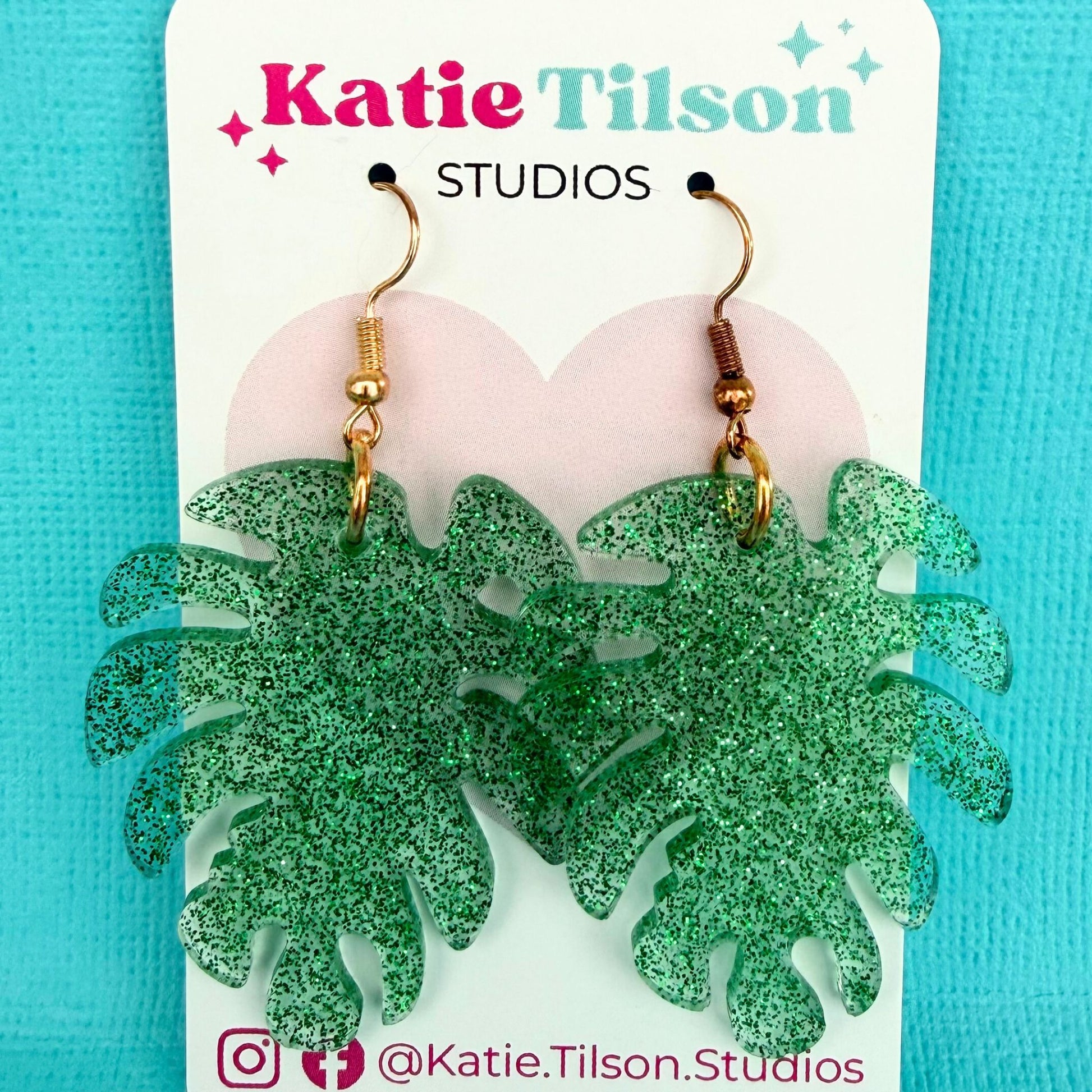 Green glittery leaf-shaped dangle earrings with gold toned hooks on a Katie Tilson Studios card against a turquoise background.