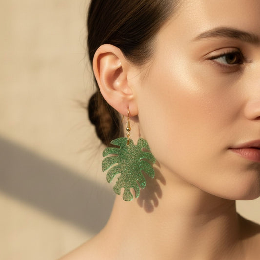 Woman wearing green monstera leaf-shaped resin dangle earrings against a neutral background.