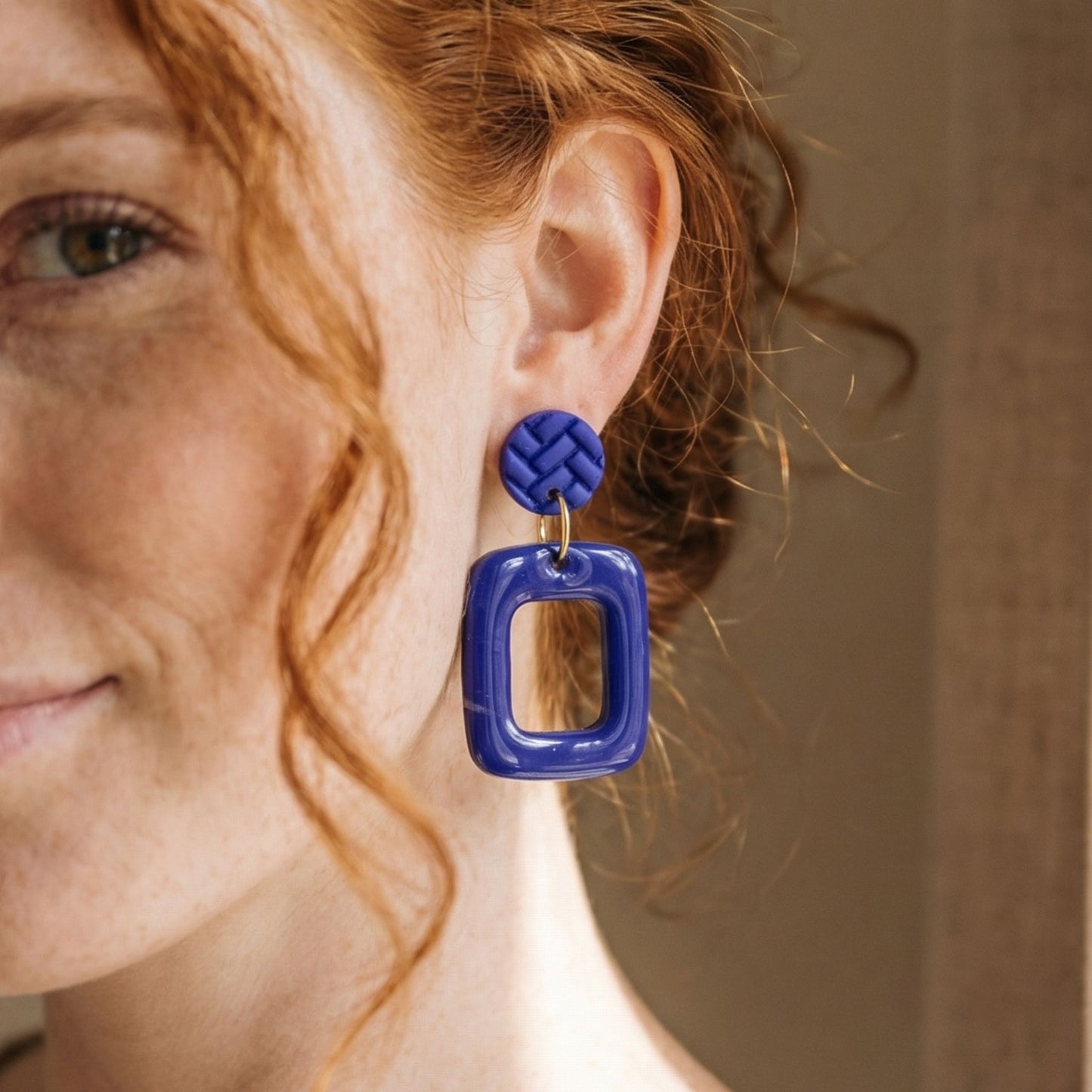 Close-up of a woman wearing cobalt blue square frame shaped drop earrings with a blurred background.