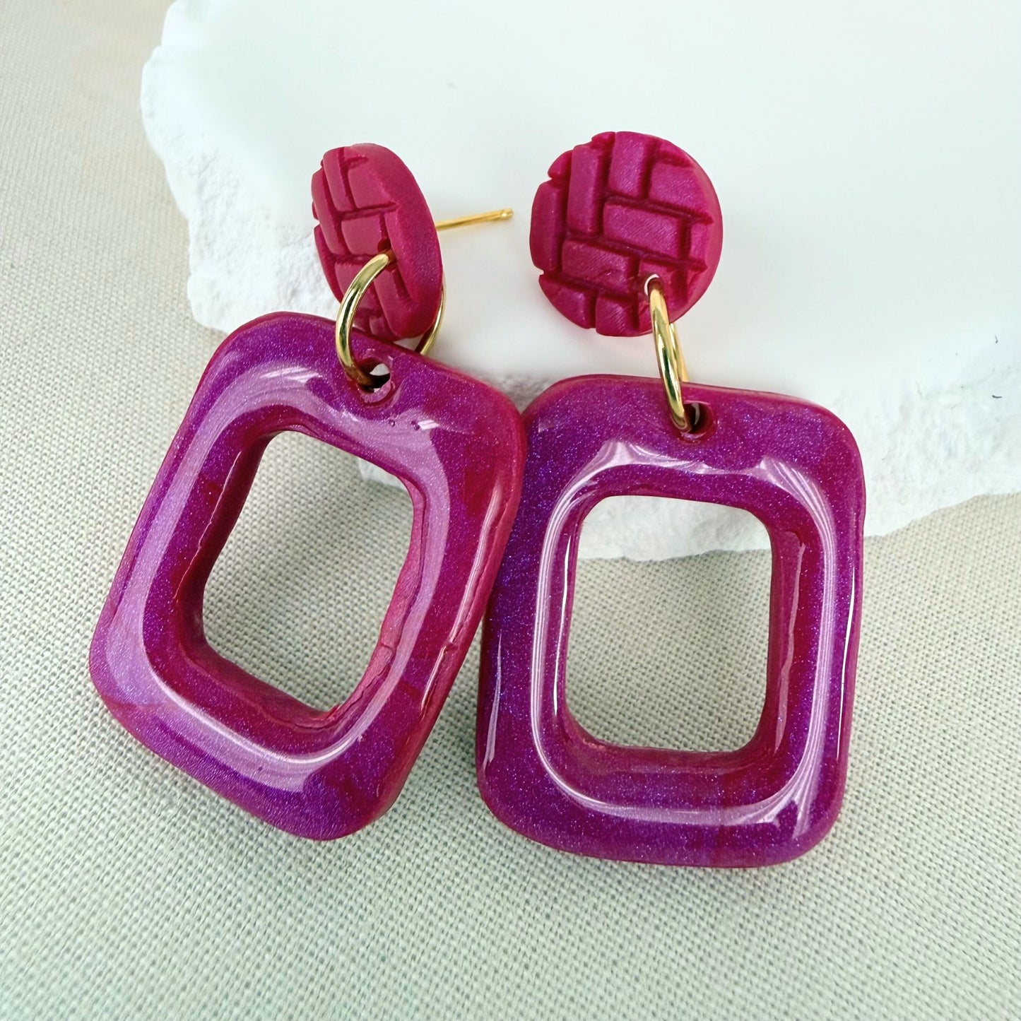 Pair of magenta frame shaped handmade drop earrings with glossy finish hanging from textured matte studs against an oat-coloured background.