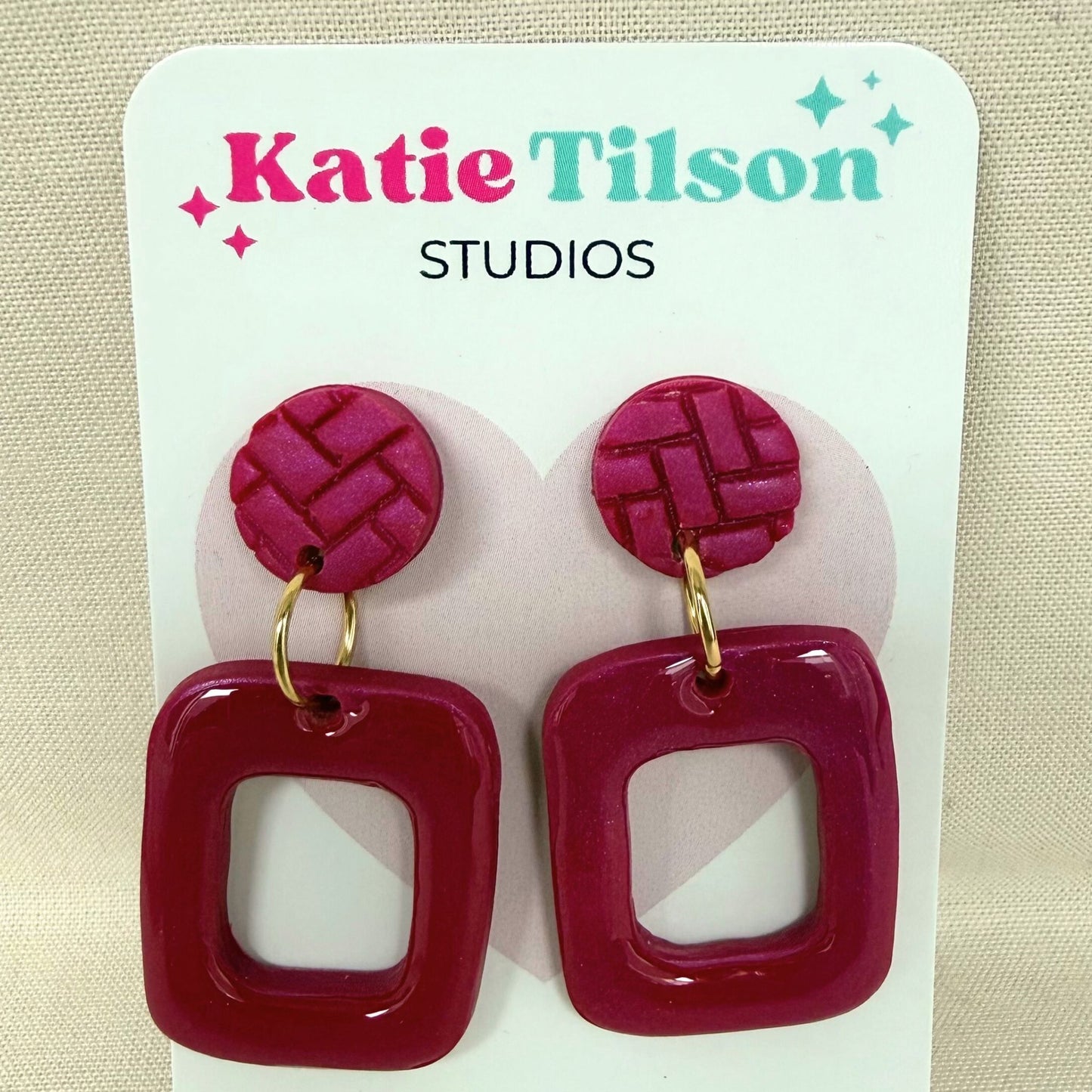 Magenta coloured frame shaped drop earrings shown on Katie Tilson Studios earring card against a neutral fabric background.