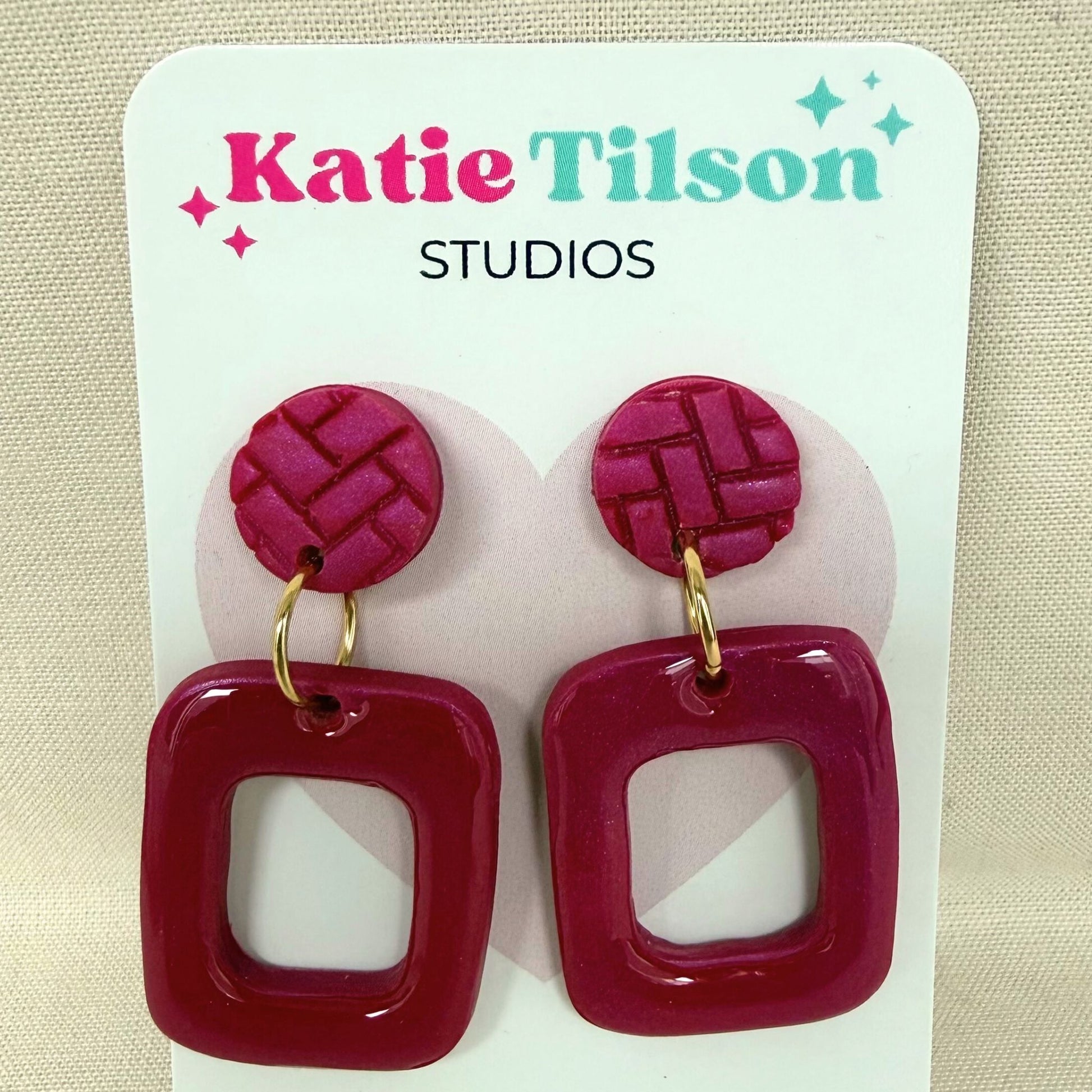 Magenta coloured frame shaped drop earrings shown on Katie Tilson Studios earring card against a neutral fabric background.