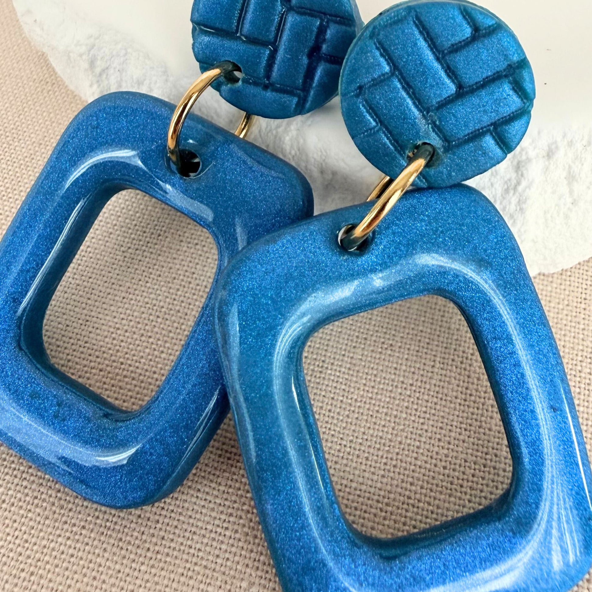 Close up of teal coloured handmade drop earrings in a rectangle frame shape with glossy finish hanging from matte studs.