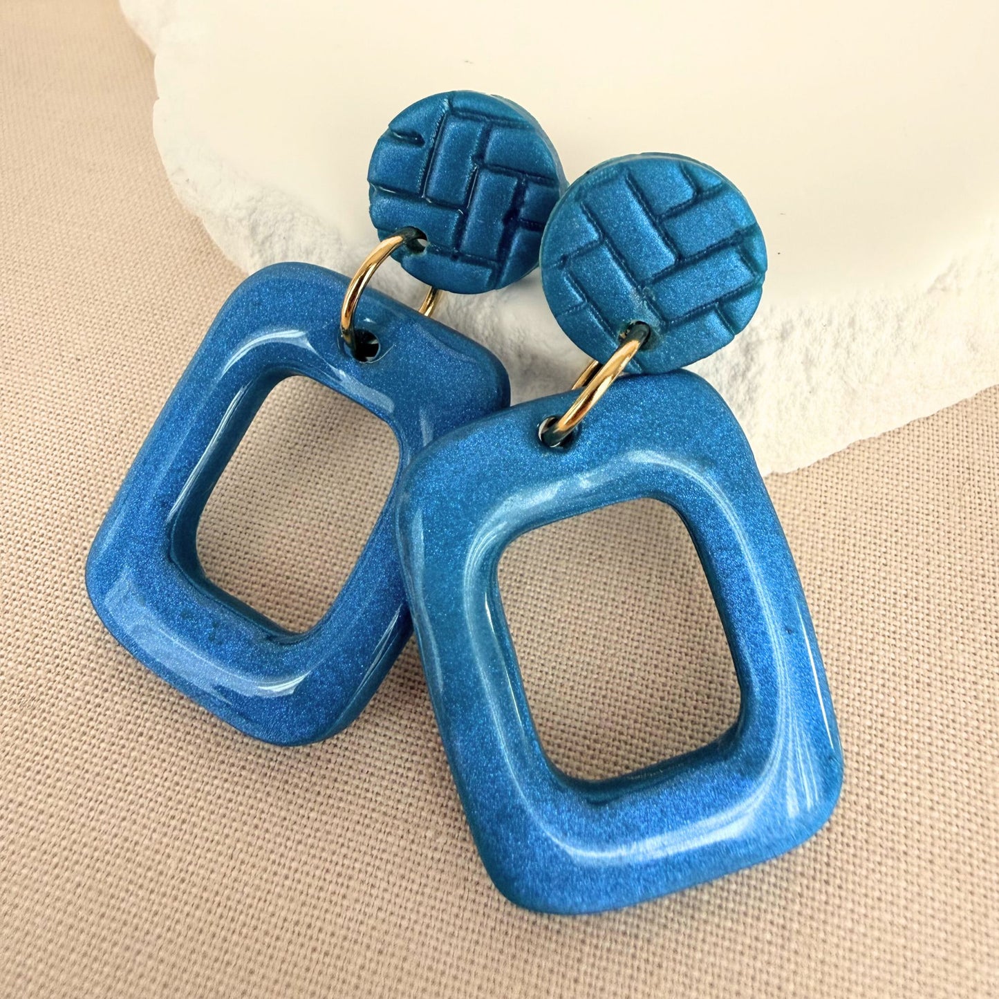 Teal coloured frame shaped handmade drop earrings with a gloss finish hanging from matte teal textured studs against an oat-coloured background.