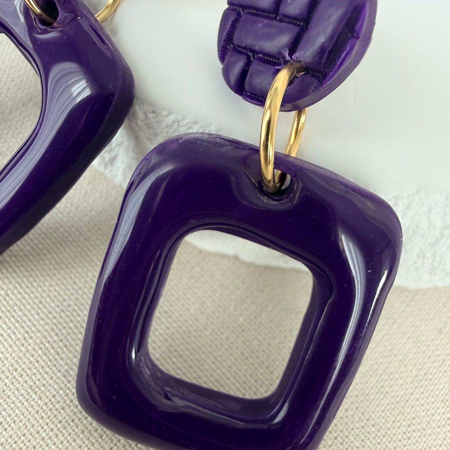 Close up of violet coloured handmade drop earrings in a rectangle frame shape with glossy finish hanging from matte studs.