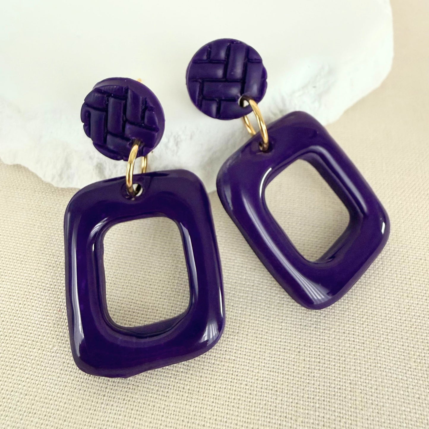 Deep violet coloured frame shaped handmade drop earrings with a high gloss finish hanging from textured violet studs on an oat-coloured background.