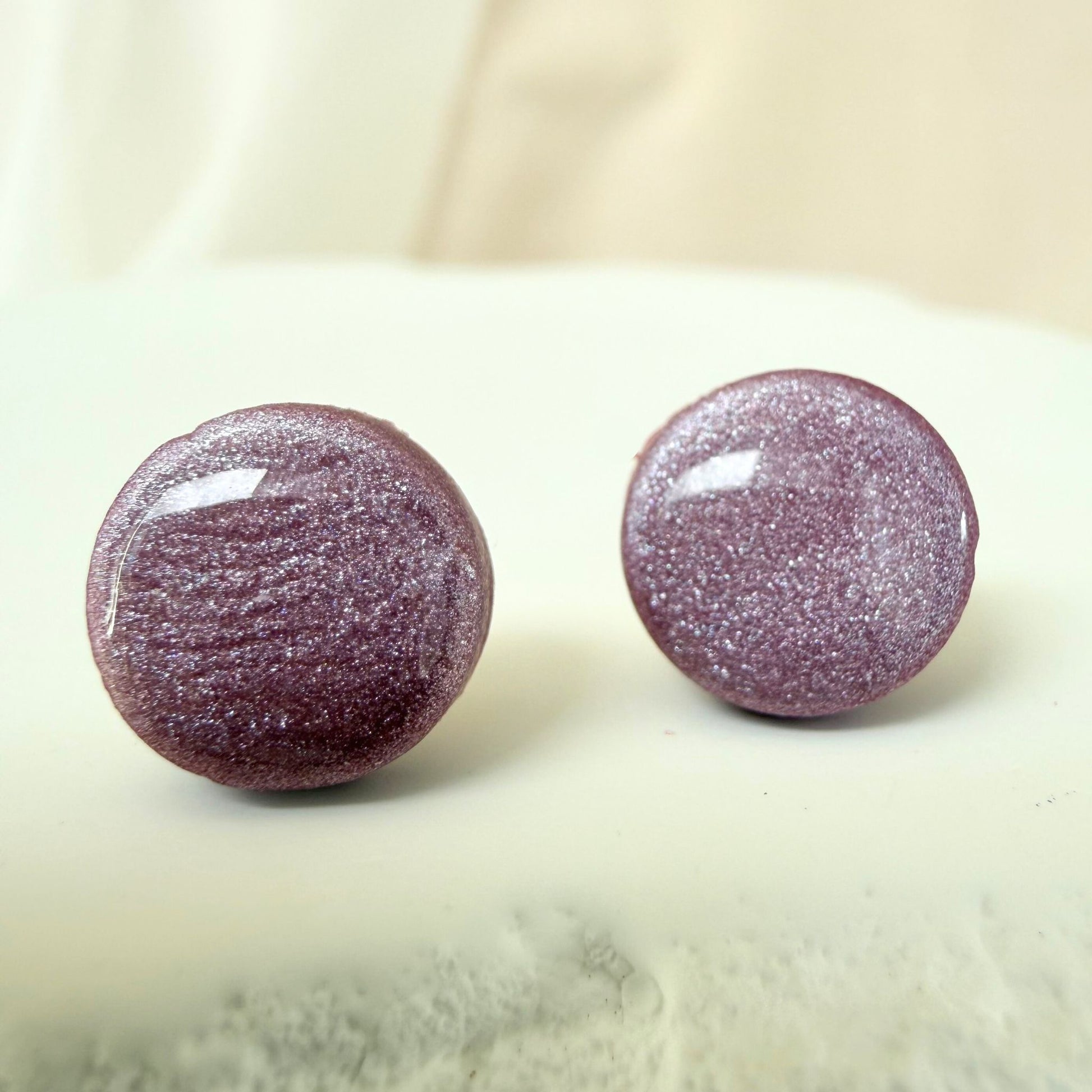 A pair of mulberry colour circle studs with shimmer and glossy resin finish resting on a white surface.