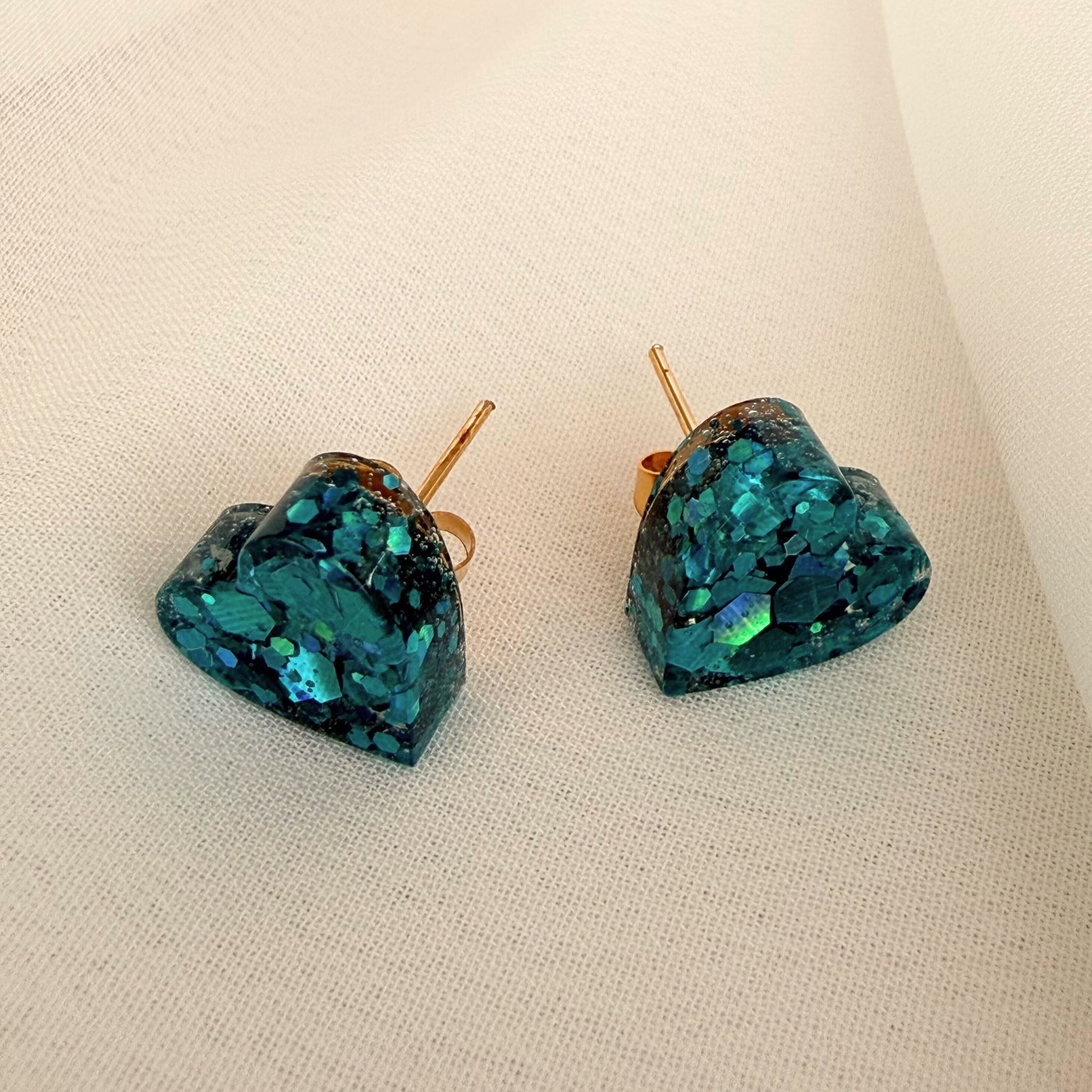 Ocean Hearts teal heart shaped stud earrings with shimmer resting on white fabric.