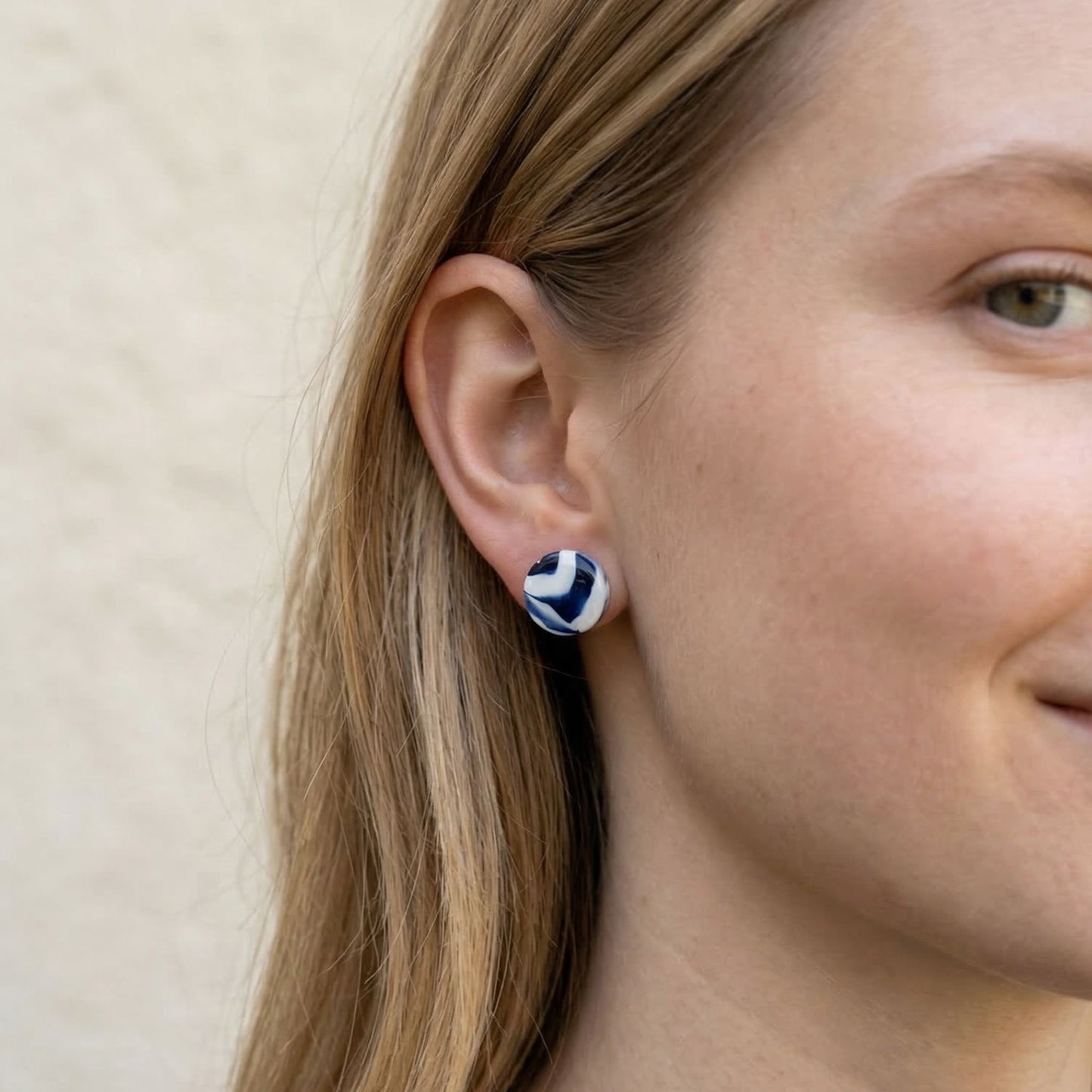 Close-up of a person wearing blue and white patterned stud earrings against a beige background.