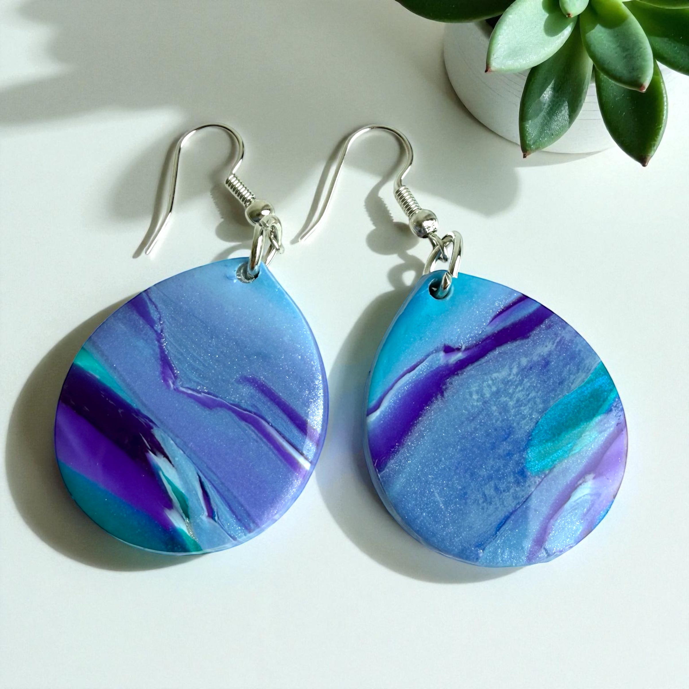 Blue and purple marbled earrings on a white surface with a plant in the background