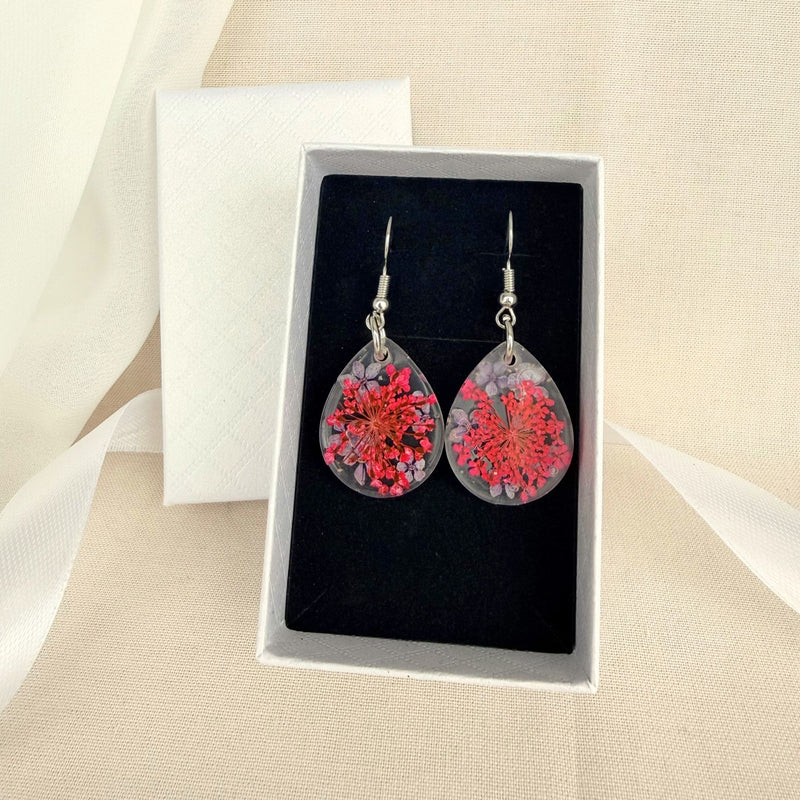 Australian handmade teardrop-shaped earrings with a pink and purple floral design in a clear resin, displayed in a box on a beige fabric background.