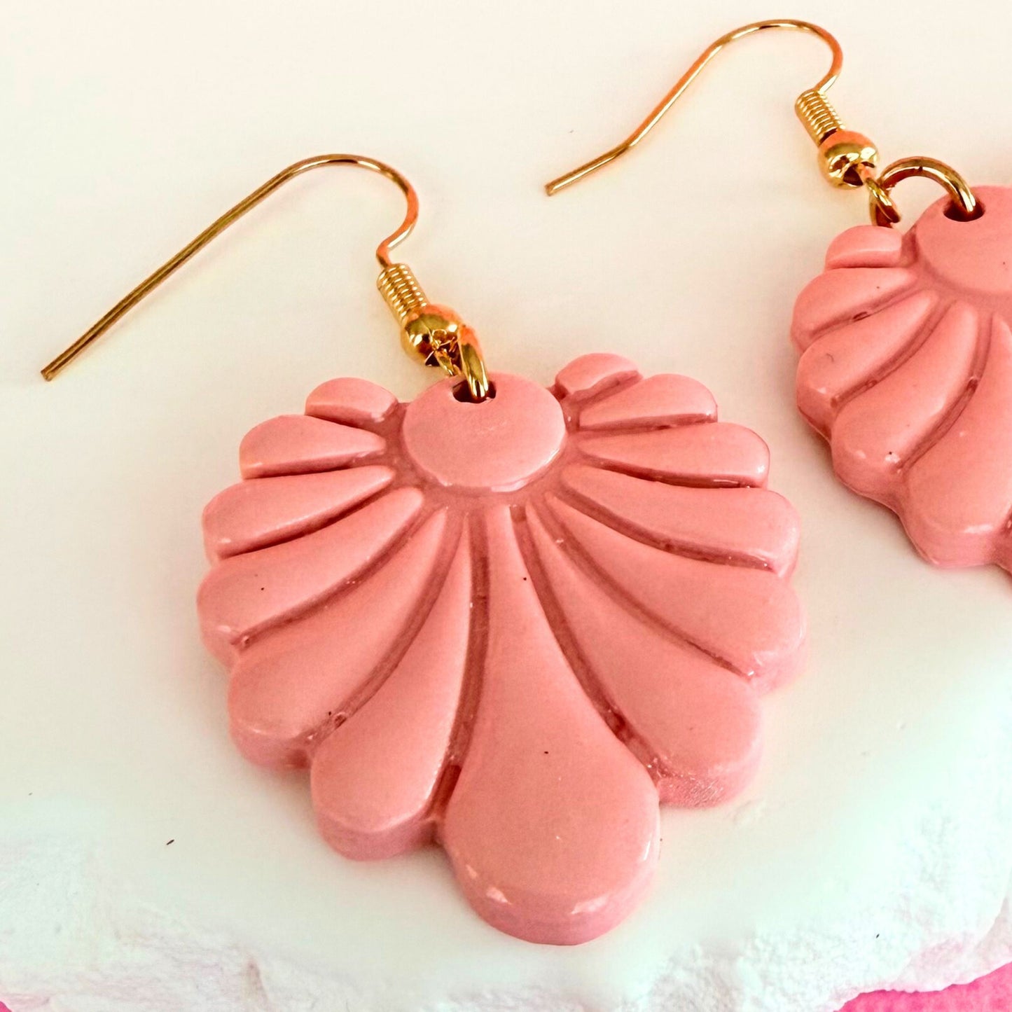 Close up of handmade dangle earrings in pink fan-shaped design with gold hooks on a light background.